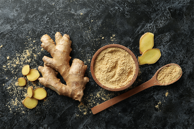 A bowl of ginger powder is placed on a black background with a ginger root and leaves placed beside the bowl.